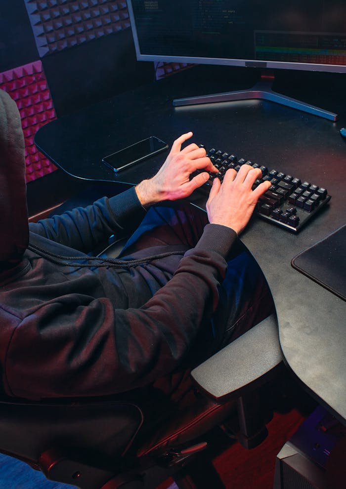 about-01 Person in hoodie typing on a keyboard in a dark room, focusing on hacking activities.